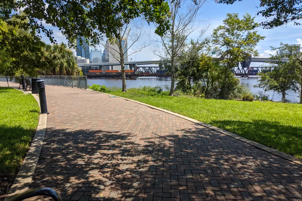 Brick path through an urban park shaded by trees with a river on the righhand side and high-rise buildings in the distance.